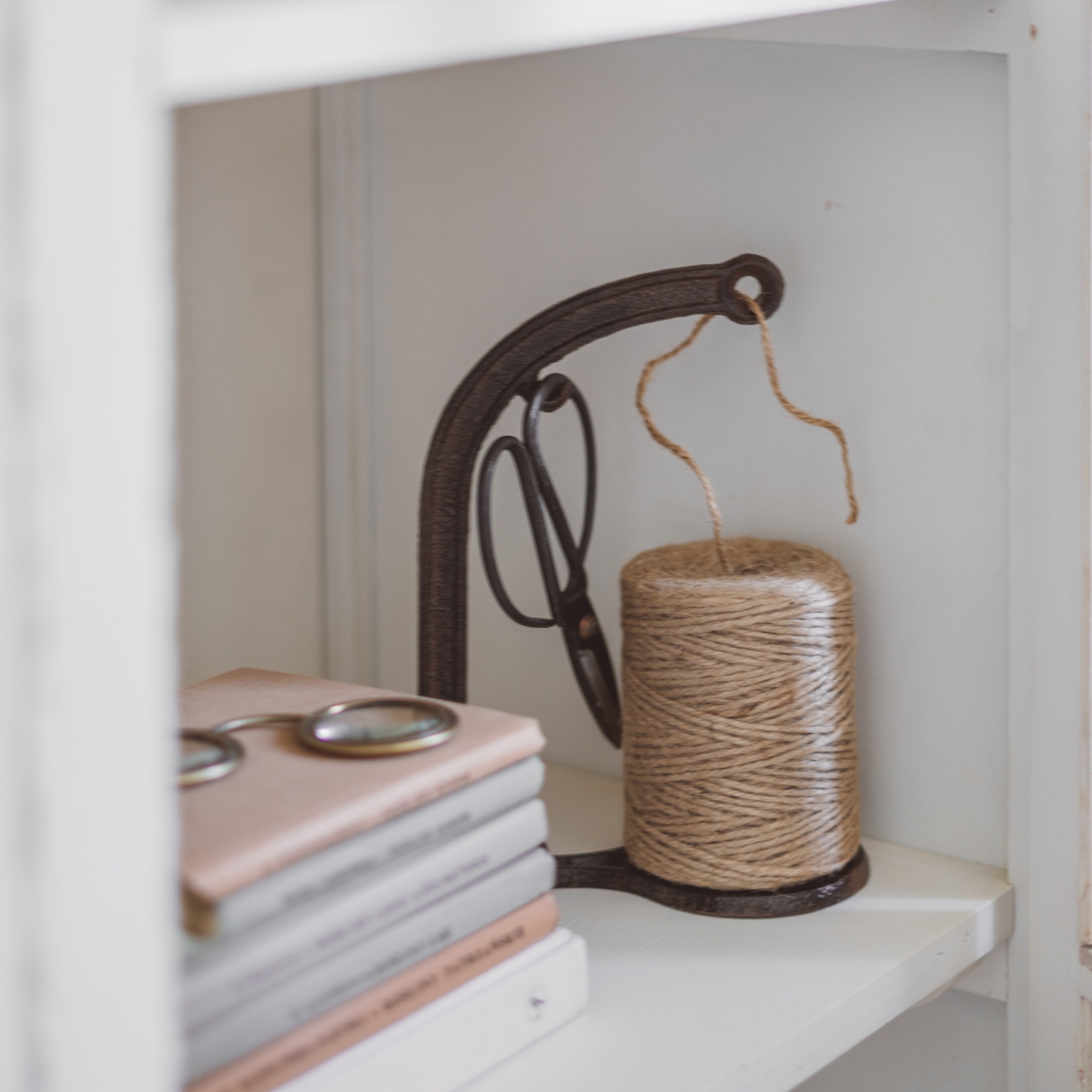 Supporto in metallo con rocchetto di spago beige e forbici su mensola bianca con libri