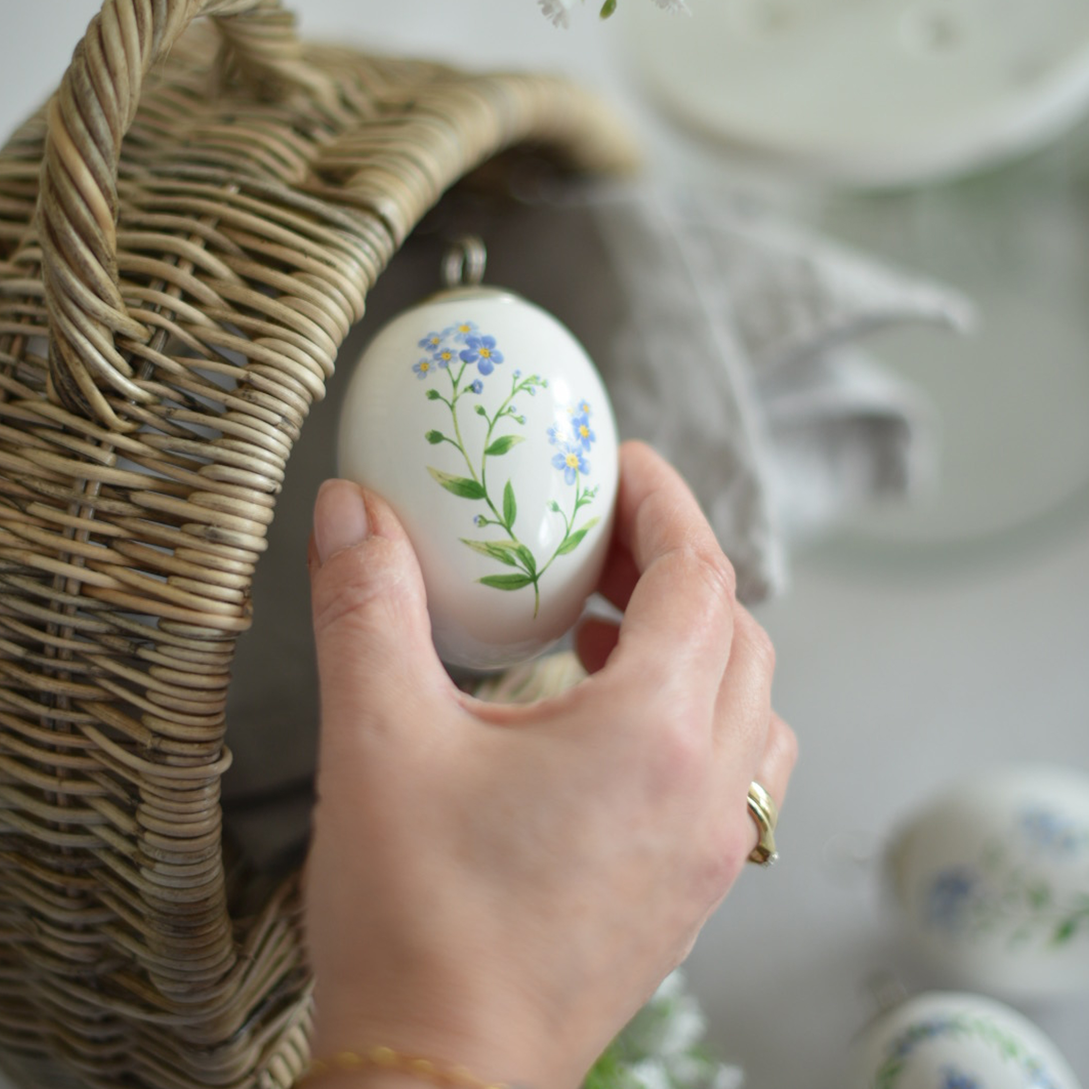 Mano che tiene decorazione pasquale in ceramica bianca con fiori blu accanto a un cesto di vimini