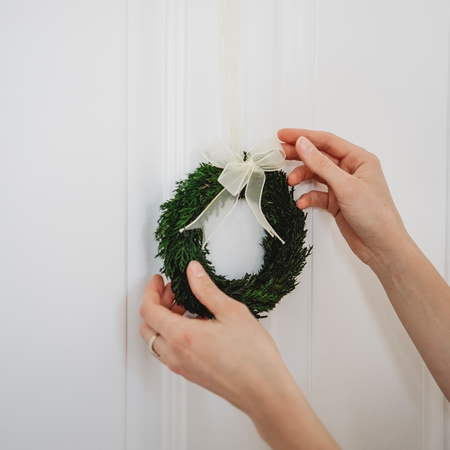 Piccola corona natalizia verde con fiocco bianco appesa a porta bianca, decorazione festiva