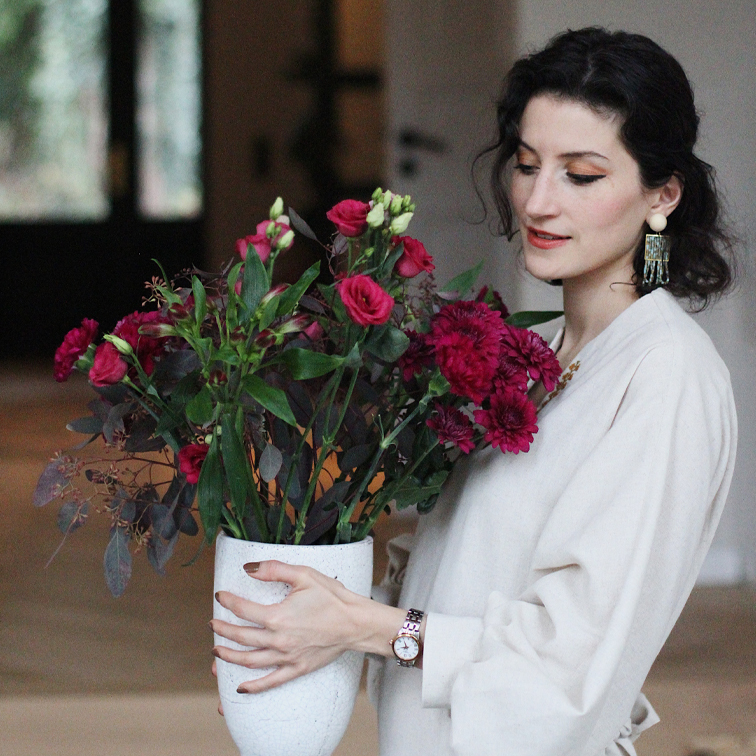 Donna con abito chiaro che tiene un vaso in ceramica bianca con fiori rossi in casa