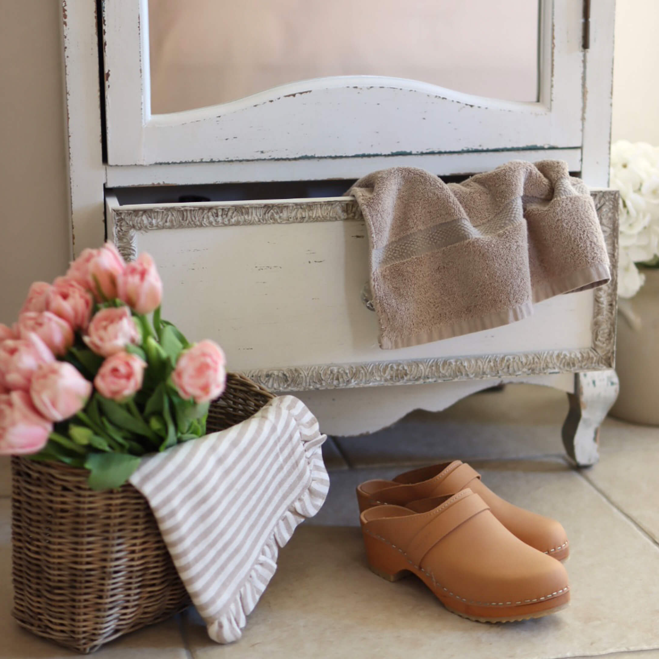 Zoccoli in pelle beige sul pavimento accanto a un cesto di vimini con fiori rosa e asciugamano