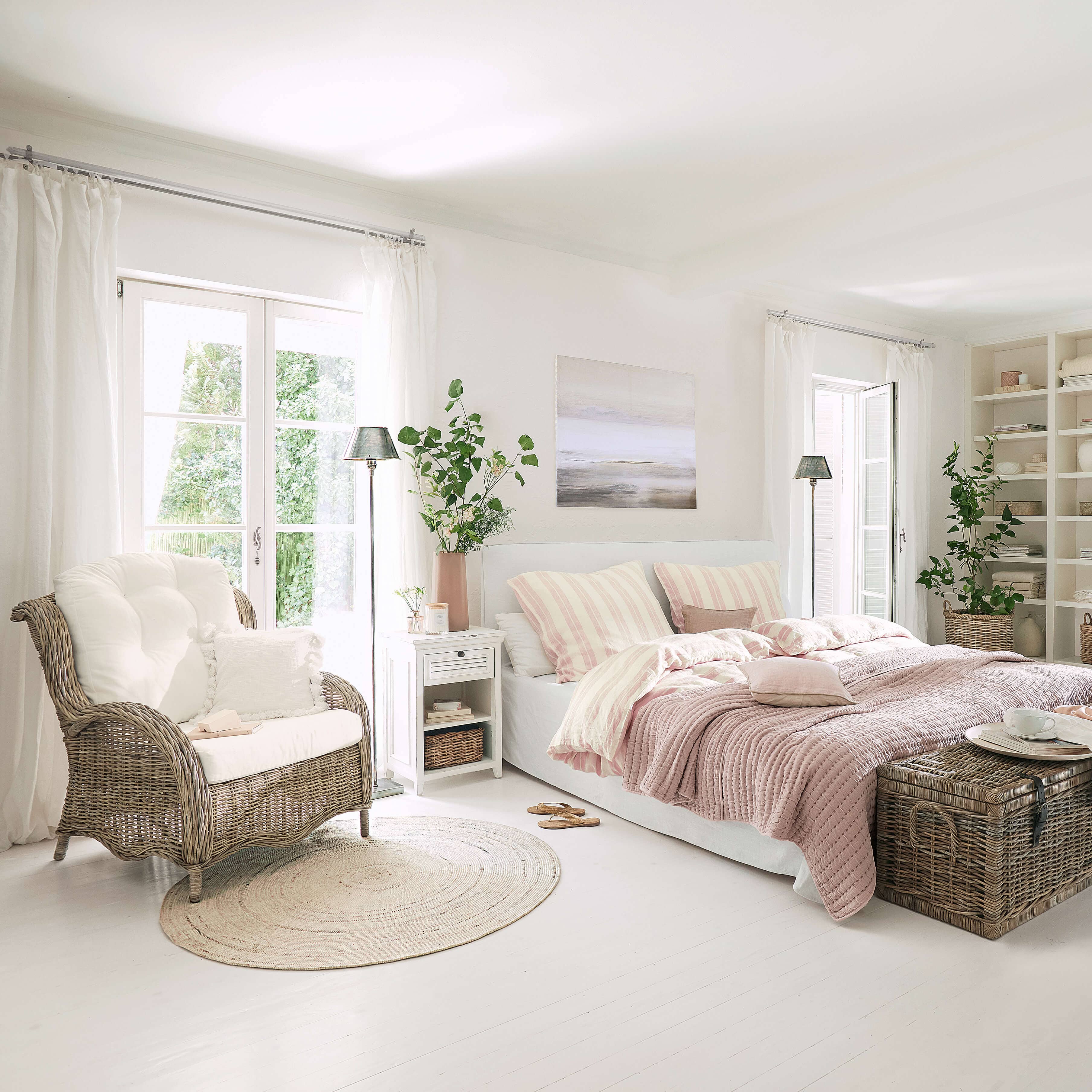 Camera luminosa con letto bianco, biancheria da letto rosa e poltrona in vimini su pavimento chiaro
