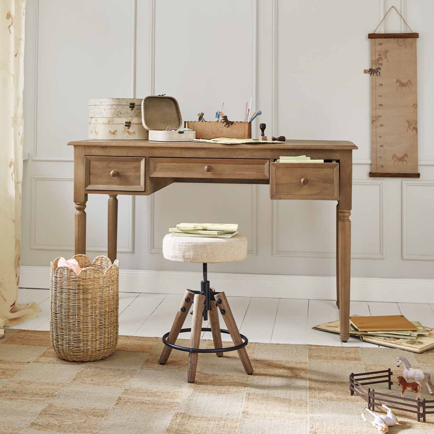 Scrivania in legno con tre cassetti e sgabello beige davanti a parete chiara con decorazioni di cavalli
